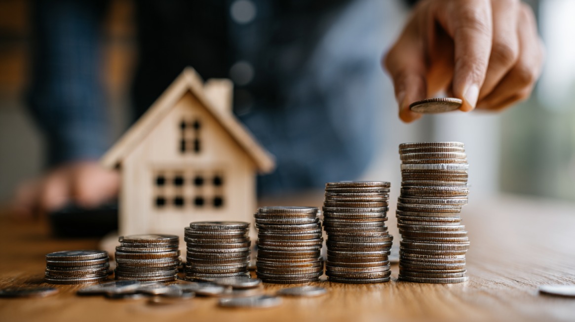 Close up of stacked coins increasing in height with a small wooden house model in the background and a hand adding a coin to one of the stacks